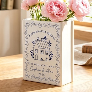 Könnte beinhalten: Eine weiße, rechteckige Keramikvase mit gerippten Seiten, verziert mit einem blauen Aufdruck eines Hauses, floralen Ranken und dem Text A NEW CHAPTER BEGINS, 724 WILLOW LANE, Sophia & Alen, 2026. Die Vase enthält einen Strauß rosa Blumen.
