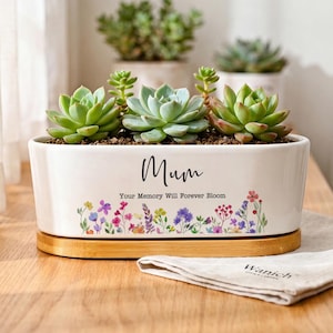 Może przedstawiać: Owalna biała ceramiczna doniczka z czarnym napisem "Mum" oraz "Your Memory Will Forever Bloom" nad kolorową grafiką kwiatową. Doniczka stoi na jasnej drewnianej podstawie i zawiera trzy zielone sukulenty o grubych liściach.