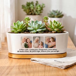 Peut inclure: Un pot de fleurs rectangulaire en céramique blanche avec une base en bambou, contenant trois plantes succulentes vertes. L'avant présente trois photos imprimées de personnes et le texte : A Nana's smile, a soothing grace. A loving touch, a warm embrace. Her kindness blooms, like flowers bright, A beacon of love, pure and light.