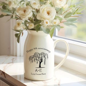 May include: A cream-colored ceramic pitcher with a handle, filled with white roses and greenery. The pitcher has a black illustration of a weeping willow tree and the text "Happy 9th anniversary" and "Love Grows Here".