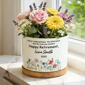 May include: A white ceramic flower pot with a floral design and the text "Happy Retirement, Luna Smith 2025." The pot is filled with a colorful arrangement of flowers, including pink roses, yellow gerbera daisies, and lavender sprigs. The pot sits on a wooden coaster.