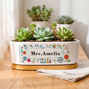 May include: A white oval ceramic planter with a bamboo base, containing three green succulents. The planter features a colourful graphic print of school supplies and the text Mrs. Amelia centred in a rectangular frame on the front side.