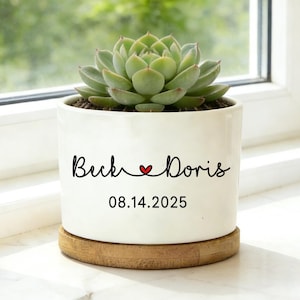 May include: A white ceramic planter with a succulent plant, sitting on a wooden coaster. The planter has the names "Buck" and "Doris" with a heart symbol, and the date "08.14.2025" in black script. The background shows a window with green foliage.