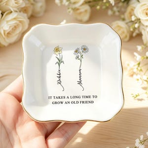 May include: A white ceramic trinket dish with a gold rim, featuring floral illustrations and the names "Debbie" and "Shannon." The dish is inscribed with the phrase "IT TAKES A LONG TIME TO GROW AN OLD FRIEND."