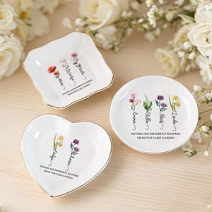 Puede incluir: Tres platos de cerámica blanca con borde dorado, cada uno con diseños florales y el texto "Sisters are different flowers from the same garden." Los platos son cuadrados, redondos y en forma de corazón.