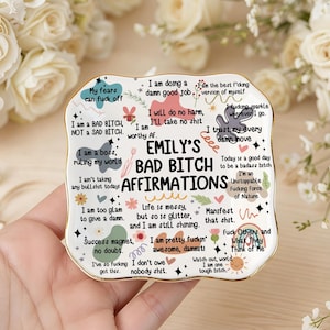 Könnte beinhalten: Ein rechteckiges Keramik-Schmucktablett mit goldenem Rand, das den Text "Emily's Bad Bitch Affirmations" in einer verspielten Schriftart trägt. Das Tablett ist mit verschiedenen Affirmationen und floralen Akzenten in verschiedenen Farben verziert.
