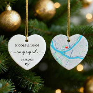 May include: Two heart-shaped Christmas ornaments hanging on a tree. One white ornament says "Nicole & Jakob engaged 05.31.2025" with a heart. The other ornament has a map design with a red heart.