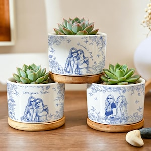 May include: Three white ceramic cylindrical planters with bamboo saucers, each containing a green succulent. The planters feature blue line-art illustrations of two people and floral patterns, with text reading "Annike & Magie" and "Love youra Bestie."