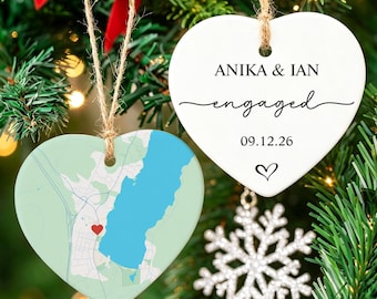 May include: Two heart-shaped ceramic ornaments hanging by twine. One ornament features a map with a red heart marker, while the other is white with black text reading ANIKA & IAN, engaged, 09.12.26, and a small heart symbol at the bottom.