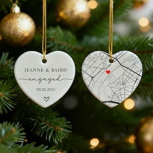 May include: Two heart-shaped Christmas ornaments hanging on a tree. One ornament reads "Jeanne & Baird engaged 09.20.2025" with a heart symbol. The other features a map design with a red heart.
