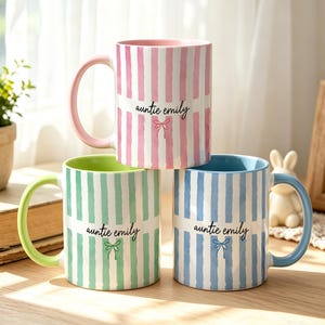 Peut inclure: Trois tasses en céramique avec des rayures verticales roses, vertes et bleues. Chaque tasse possède une anse et un intérieur de couleur assortie. Le texte "auntie emily" est imprimé en noir sur le devant, avec une petite icône de nœud.