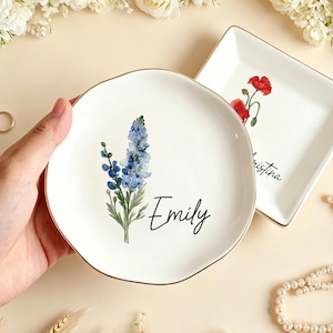 Puede incluir: Dos platos de cerámica blancos para anillos con borde dorado. Uno es redondo con un diseño floral azul y el nombre "Emily". El otro es cuadrado con un diseño floral rojo y el nombre "Kristina". También hay un anillo de oro y un collar de perlas.