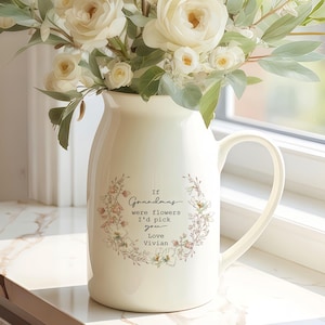 Puede incluir: Una jarra de cerámica color crema con asa, llena de rosas blancas y vegetación. La jarra tiene un diseño floral y el texto "If grandmas were flowers I'd pick you. Love Vivian."