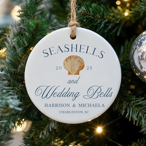 Może przedstawiać: Biała ceramiczna ozdoba choinkowa z napisem "Seashells and Wedding Bells" w granatowym skrypcie. Grafika muszli jest wyśrodkowana nad tekstem. Ozdoba wisi na choince ze srebrnymi bombkami.