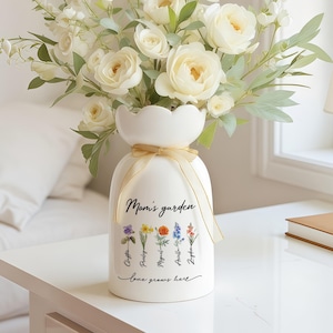 Könnte beinhalten: Eine weiße Keramikvase mit gewelltem Rand, gefüllt mit weißen Rosen und Grünzeug. Die Vase hat ein goldenes Band um sich gebunden und trägt den Text "Mom's garden" mit Blumenillustrationen und den Worten "love grows here".