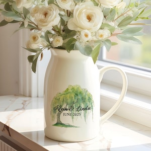 Puede incluir: Una jarra de cerámica color crema con asa, llena de rosas blancas y vegetación. La jarra tiene un diseño de sauce verde con los nombres "Klein & Linda" y la fecha "JUNIO 2023". El arreglo está sobre un alféizar blanco.