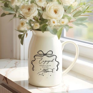 May include: A cream-colored ceramic pitcher vase filled with a bouquet of white roses and green eucalyptus foliage. The front of the pitcher features a black line-art graphic of a bow, the word Engaged, the names Vivian & Oscar, and 12.16.2025.
