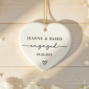 May include: A white heart-shaped ornament with the words "JEANNE & BAIRD engaged 09.20.2025" in black script. A small heart icon is below the date. The ornament hangs from a gold string, surrounded by white flowers.