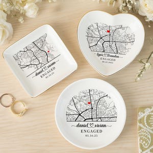 Puede incluir: Tres platos blancos de cerámica para anillos con borde dorado, cada uno con un diseño de mapa en blanco y negro con un corazón rojo. Los platos son cuadrados, en forma de corazón y redondos, con las palabras "daniel & vivian ENGAGED 05.16.25" y "Lysie & Brandon ENGAGED 08.22.2025".