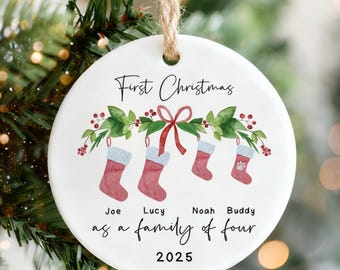 May include: A white, round Christmas ornament with the text "First Christmas" and "as a family of four 2025". It features four hanging stockings with names Joe, Lucy, Noah, and Buddy. The ornament hangs from a jute string.