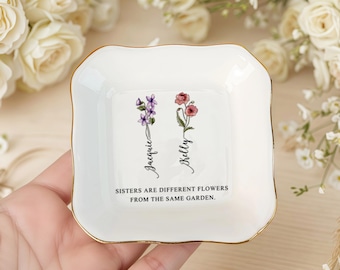May include: A white ceramic trinket dish with a gold rim. The dish features floral illustrations and the names "Jacquie" and "Kelly" in script. The text "Sisters are different flowers from the same garden" is also present.