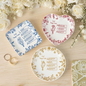 May include: Three decorative ceramic plates with floral designs and inspirational text. One is square with blue accents, one heart-shaped with red accents, and one round with gold accents. The plates are arranged on a light wood surface with flowers.