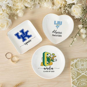 May include: Three personalized ceramic ring dishes with gold trim. One square dish features a blue logo and the name "ryann". A heart-shaped dish has a logo and the name "Alyssa". A round dish displays a cartoon duck and the name "amelia".