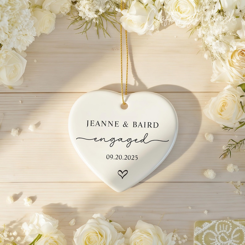 May include: A white heart-shaped ornament with the words "JEANNE & BAIRD engaged 09.20.2025" in black script. A small heart icon is below the date. The ornament hangs from a gold string, surrounded by white flowers.