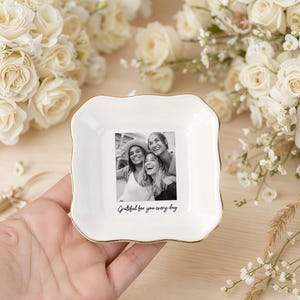 Peut inclure: Un vide-poche en c&eacute;ramique blanche avec un bord dor&eacute;, pr&eacute;sentant une photo en noir et blanc de trois personnes souriantes. Le plat porte l'inscription "Grateful for you every day". Le fond comprend des roses blanches et de la verdure.