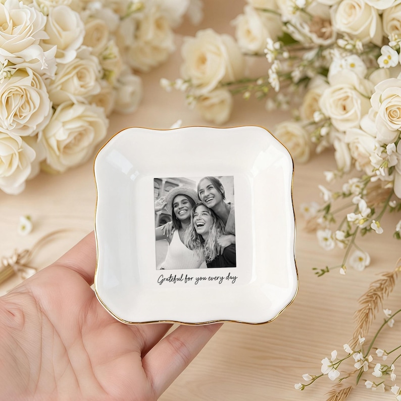 Peut inclure: Un vide-poche en c&eacute;ramique blanche avec un bord dor&eacute;, pr&eacute;sentant une photo en noir et blanc de trois personnes souriantes. Le plat porte l'inscription "Grateful for you every day". Le fond comprend des roses blanches et de la verdure.