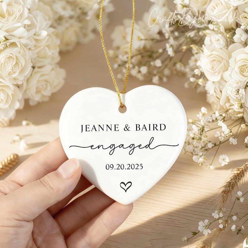 May include: A white heart-shaped ornament with gold hanging string. The ornament has the names "JEANNE & BAIRD" and the word "engaged" in script. The date "09.20.2025" and a heart symbol are also printed on the ornament. The ornament is held by a hand.