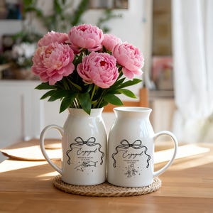 May include: Two white ceramic pitchers with handles, each featuring a black line drawing of a bow, the word Engaged, and the names Vivian & Oscar with the date 12.14.2023. One pitcher shows hands touching, the other shows two glasses clinking.
