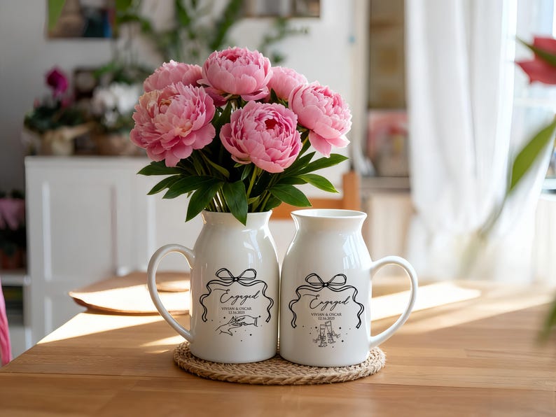 May include: Two white ceramic pitchers with handles, each featuring a black line drawing of a bow, the word Engaged, and the names Vivian & Oscar with the date 12.14.2023. One pitcher shows hands touching, the other shows two glasses clinking.