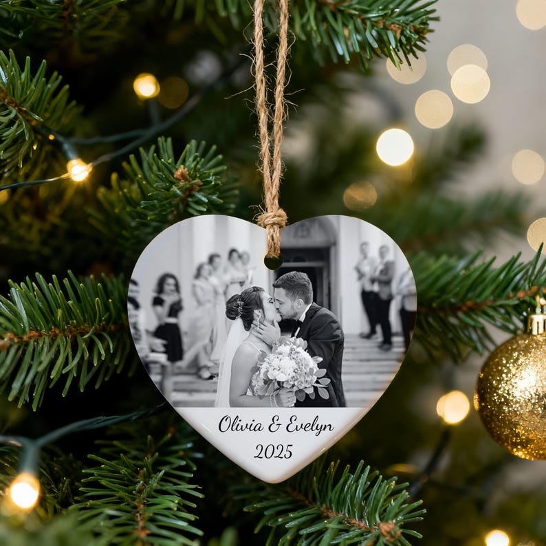 Peut inclure: Ornement de No&euml;l en forme de c&oelig;ur avec une photo de mariage en noir et blanc. L'ornement a un bord blanc et une ficelle de jute pour l'accrocher. Le texte sur l'ornement indique "Olivia & Evelyn 2025". L'ornement est accroch&eacute; &agrave; un sapin de No&euml;l.