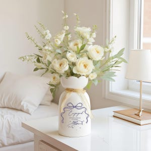 May include: A white ceramic vase with a scalloped rim and a gold ribbon tied around the neck. The vase features a purple graphic of a bow, the word "Engaged," the names "Vivian & Oscar," the date "12.16.2021," and an illustration of two hands.