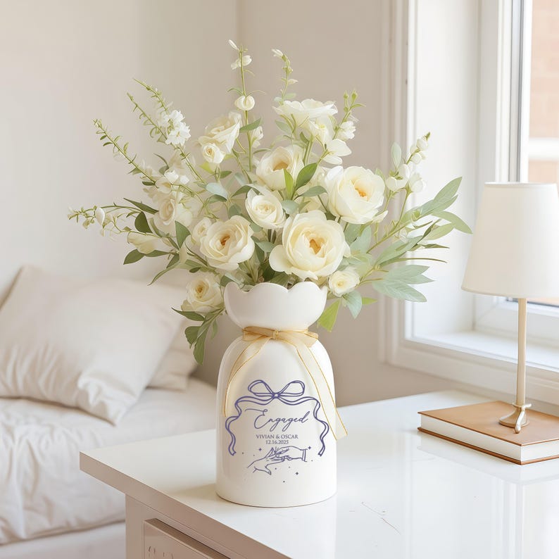 May include: A white ceramic vase with a scalloped rim and a gold ribbon tied around the neck. The vase features a purple graphic of a bow, the word "Engaged," the names "Vivian & Oscar," the date "12.16.2021," and an illustration of two hands.