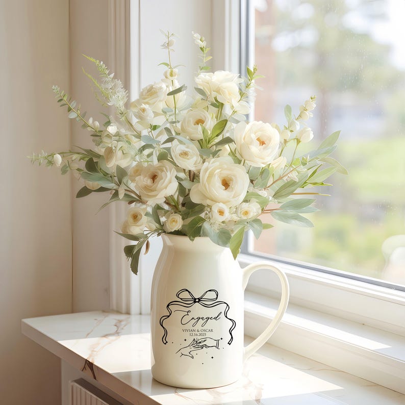 May include: A cream-colored ceramic pitcher vase filled with a bouquet of white roses and green eucalyptus foliage. The front of the pitcher features a black line-art graphic of a bow, the word Engaged, the names Vivian & Oscar, and 12.16.2025.