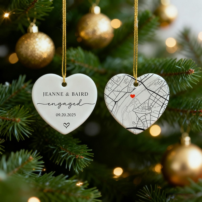 May include: Two heart-shaped Christmas ornaments hanging on a tree. One ornament reads "Jeanne & Baird engaged 09.20.2025" with a heart symbol. The other features a map design with a red heart.