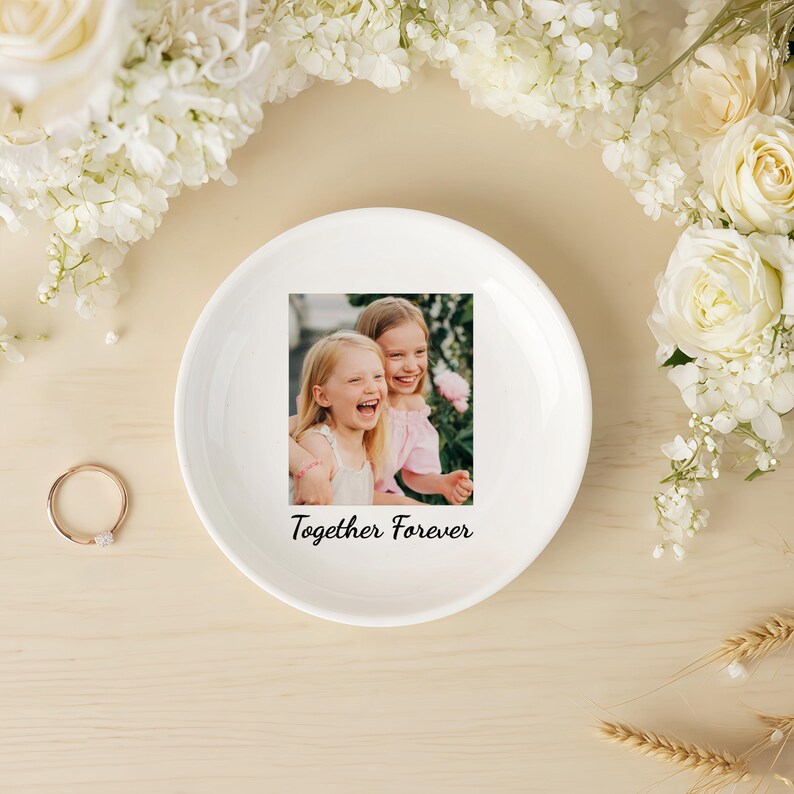 Peut inclure: Un vide-poche en c&eacute;ramique blanche avec une photo de deux filles riant et le texte "Together Forever". Le plat est entour&eacute; de fleurs blanches et d'une bague en or. Le fond est de couleur beige clair.