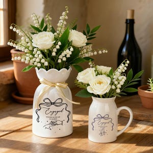 May include: Two white ceramic vessels, a vase and a pitcher, each featuring a black line drawing of a bow, the text "Engaged," names, and a date. The vessels are filled with white roses and lily of the valley flowers, accented with a gold ribbon.