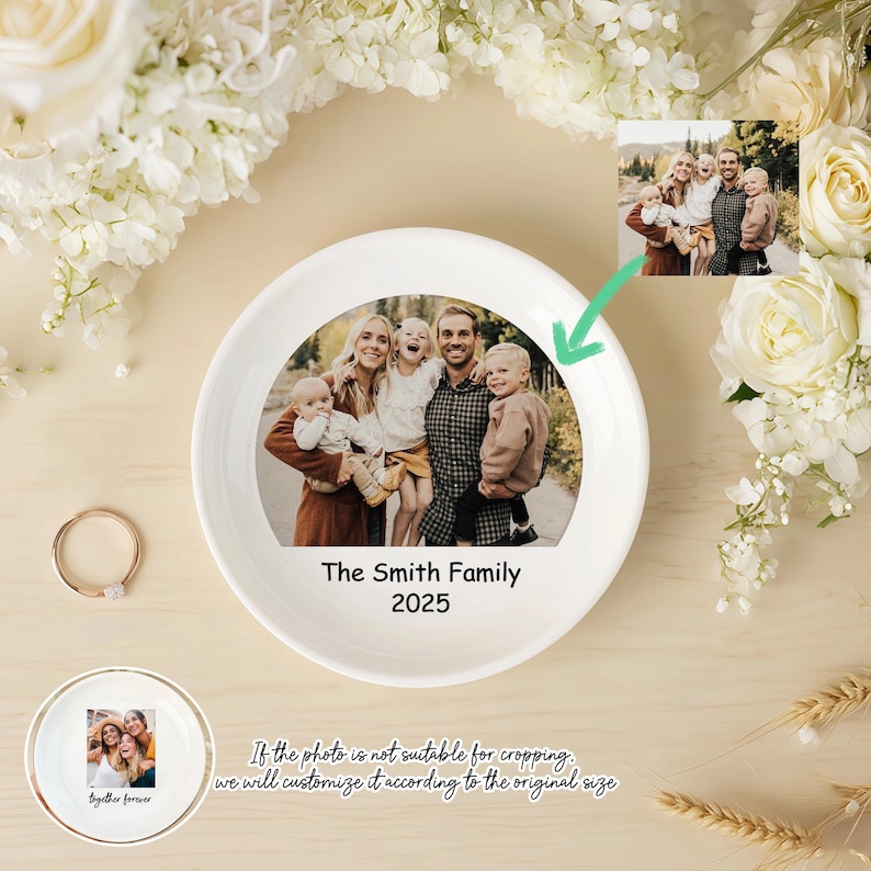 Peut inclure: Un vide-poche en c&eacute;ramique blanche avec une photo de famille et le texte "The Smith Family 2025". Une petite bague et une seconde coupelle avec une photo sont &eacute;galement visibles. Le plat est entour&eacute; de fleurs blanches.