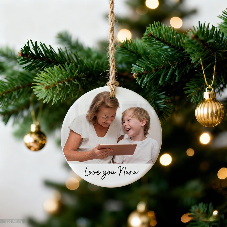 Peut inclure: Une d&eacute;coration de No&euml;l ronde en c&eacute;ramique blanche avec une photo d'une femme et d'un enfant regardant une tablette. Le texte "Love you Nana" est imprim&eacute; en bas. L'ornement est suspendu &agrave; une ficelle de jute sur un sapin de No&euml;l.