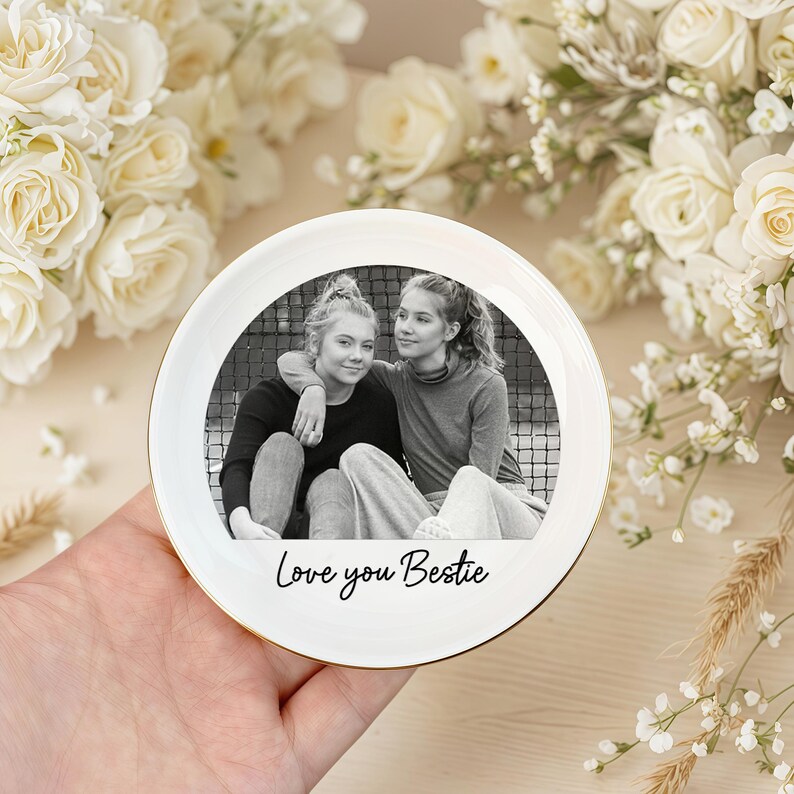 Peut inclure: Une assiette en c&eacute;ramique blanche avec un bord dor&eacute;, orn&eacute;e d'une photo en noir et blanc de deux jeunes femmes enlac&eacute;es. L'assiette porte l'inscription "Love you Bestie". L'assiette est tenue dans une main, entour&eacute;e de fleurs blanches.