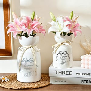 May include: Two white ceramic vases with scalloped rims, each tied with a cream ribbon. The vases feature black printed text reading Engaged, names, dates, and illustrations of hearts or champagne flutes, filled with pink and white lily flowers.