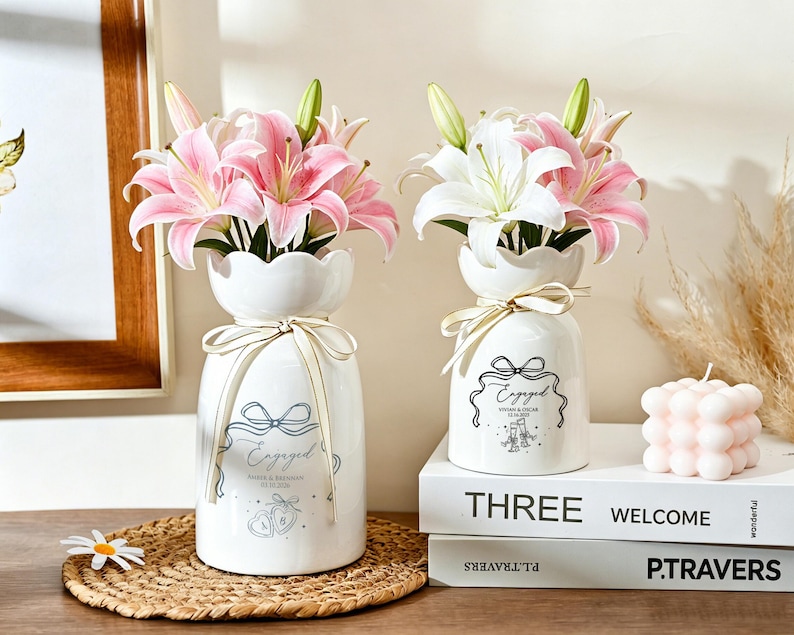 May include: Two white ceramic vases with scalloped rims, each tied with a cream ribbon. The vases feature black printed text reading Engaged, names, dates, and illustrations of hearts or champagne flutes, filled with pink and white lily flowers.