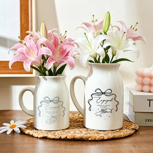 May include: Two white ceramic milk-style pitchers with handles, each featuring a black line-art graphic of a ribbon, the word "Engaged," names, and a date. The left pitcher holds pink lilies, and the right pitcher holds white lilies.