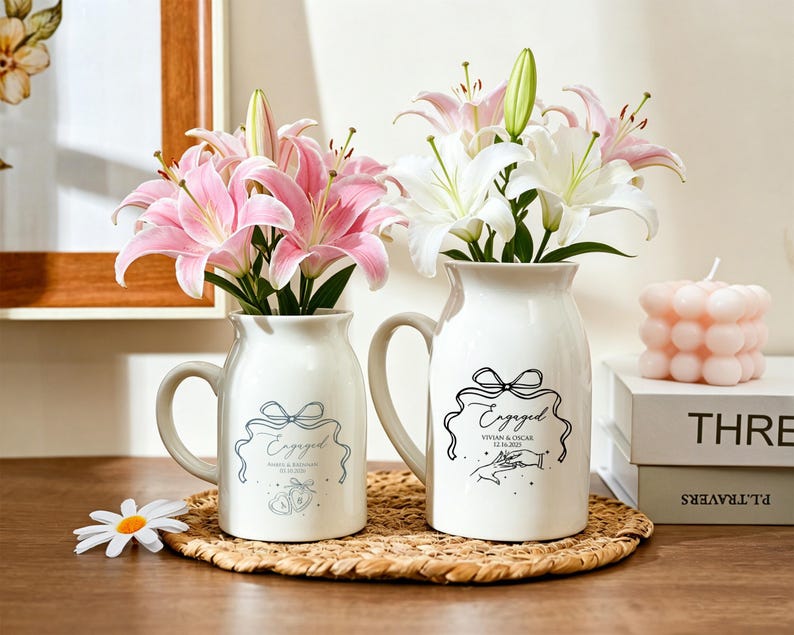 May include: Two white ceramic milk-style pitchers with handles, each featuring a black line-art graphic of a ribbon, the word "Engaged," names, and a date. The left pitcher holds pink lilies, and the right pitcher holds white lilies.