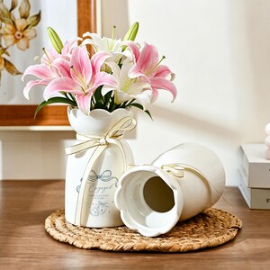 May include: Two white ceramic vases with scalloped rims and cream-colored ribbons tied around the necks. One vase is upright, filled with pink and white lilies, and features the text Engaged with a bow and ring graphic. The other vase lies on its side.