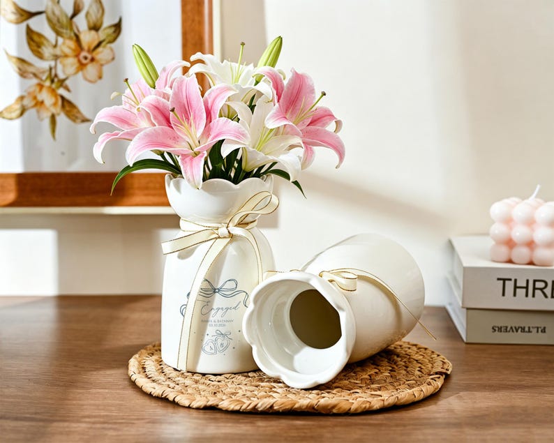 May include: Two white ceramic vases with scalloped rims and cream-colored ribbons tied around the necks. One vase is upright, filled with pink and white lilies, and features the text Engaged with a bow and ring graphic. The other vase lies on its side.