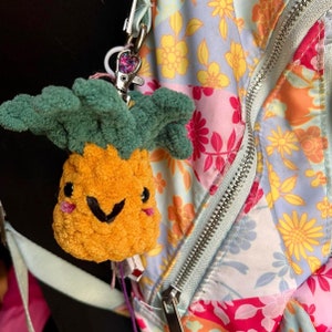 Op de afbeelding: Een gele ananasvormige pluche sleutelhanger met een groene, bladerige bovenkant en een glimlachend gezicht. De sleutelhanger is bevestigd aan een zilveren rits aan een tas met bloemenprint.